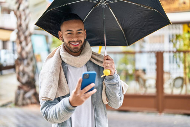 African american man using smartphone holding umbrella at street - Zdjęcie, obraz