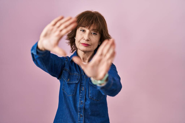 Donna di mezza età in piedi su sfondo rosa facendo cornice con mani palme e dita, prospettiva fotocamera  - Foto, immagini