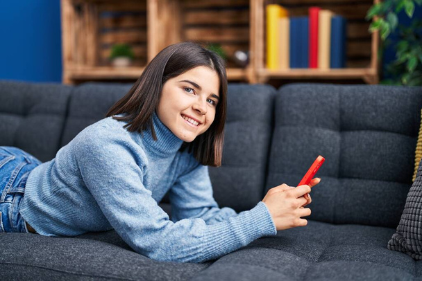 Молодая красивая латиноамериканка с помощью смартфона лежит дома на диване - Фото, изображение
