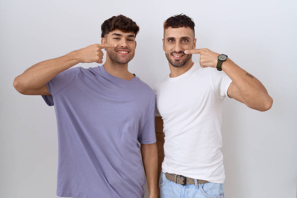 Couple homosexuel gay debout sur fond blanc pointant avec le doigt de la main vers le visage et le nez, souriant gai. concept de beauté  - Photo, image