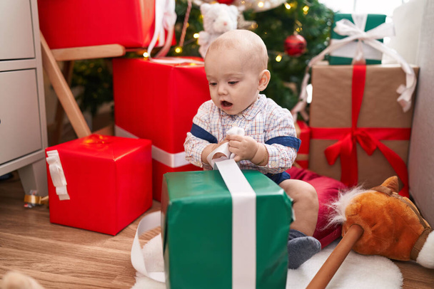 Schattige Kaukasische baby uitpakken geschenk zittend op de vloer door kerstboom thuis - Foto, afbeelding