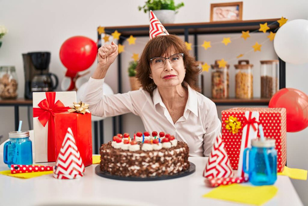 Женщина средних лет празднует день рождения, держа в руках большой шоколадный торт, раздраженный и разочарованный, крича от гнева, крича сумасшедшие от злости и подняв руку  - Фото, изображение
