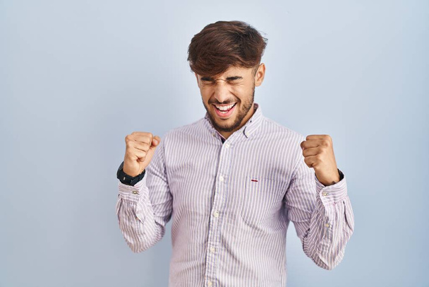 Arab man with beard standing over blue background very happy and excited doing winner gesture with arms raised, smiling and screaming for success. celebration concept.  - Photo, Image