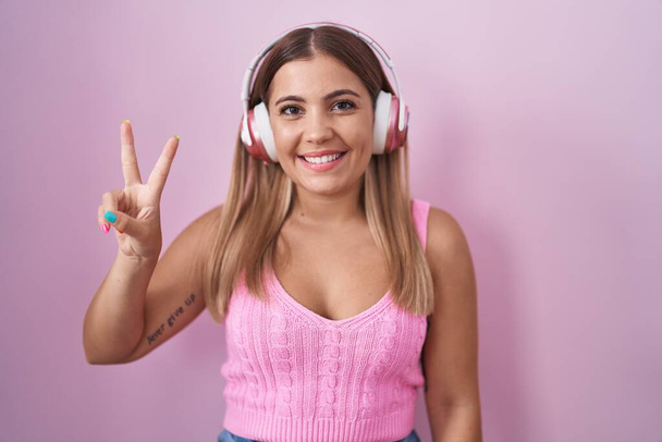 Mujer rubia joven escuchando música usando auriculares mostrando y apuntando hacia arriba con los dedos número dos mientras sonríe confiada y feliz.  - Foto, Imagen