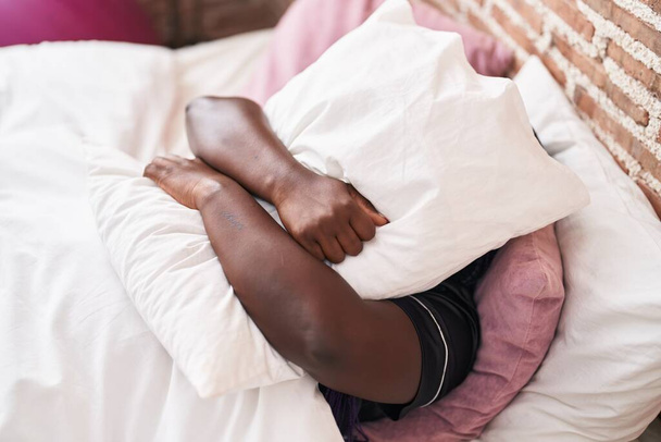 Αφροαμερικανή γυναίκα που καλύπτει το πρόσωπο με μαξιλάρι ξαπλωμένη στο κρεβάτι στο υπνοδωμάτιο - Φωτογραφία, εικόνα