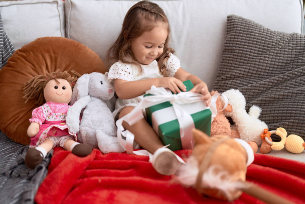 Rozkošná hispánská dívka drží dárek sedí na pohovce doma - Fotografie, Obrázek