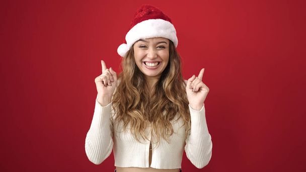 クリスマスの帽子をかぶった若い美しいヒスパニックの女性は,孤立した赤い背景の上に育った指で指を指します. - 写真・画像