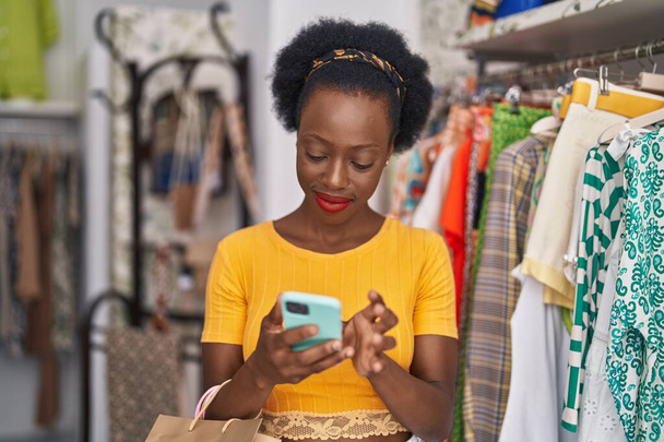 Afrikaanse Amerikaanse vrouw klant houden boodschappentassen met behulp van smartphone in de kledingwinkel - Foto, afbeelding