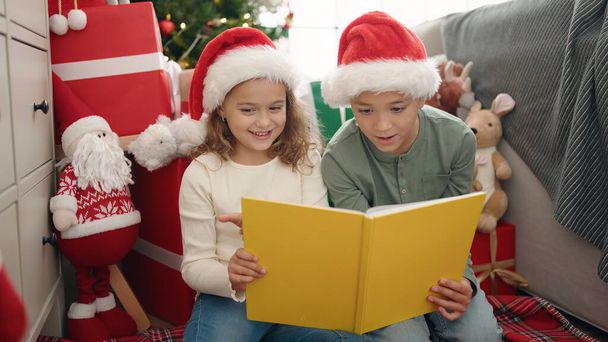 Δύο παιδιά διαβάζουν το βιβλίο κάθεται στο πάτωμα από το χριστουγεννιάτικο δέντρο στο σπίτι - Φωτογραφία, εικόνα