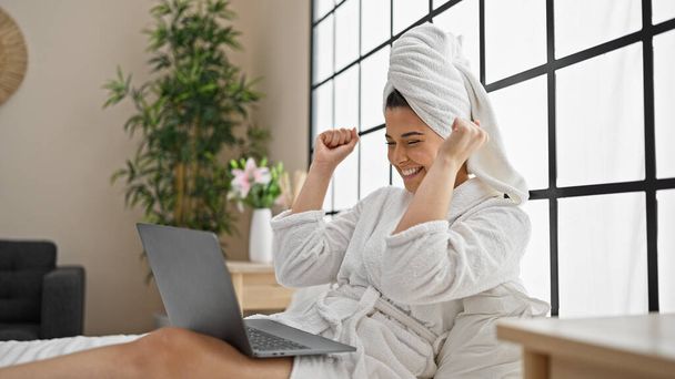 Jovem bela mulher hispânica vestindo roupão de banho usando laptop celebrando no quarto - Foto, Imagem