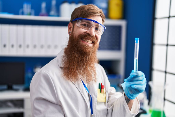 Mladý ryšavý muž vědec s úsměvem jistý držení zkumavky v laboratoři - Fotografie, Obrázek