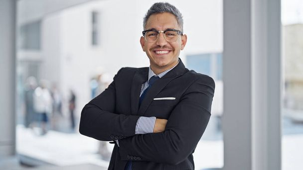 Молодий іспаномовний чоловік бізнесмен посміхається впевнено стоячи зі зброєю перетнув жест в офісі
 - Фото, зображення