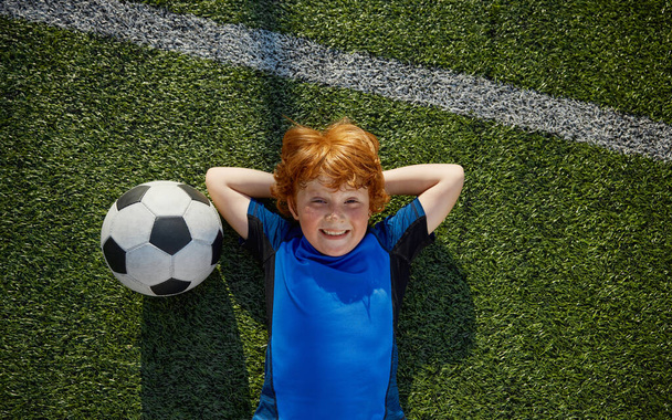 Relajado niño niño pequeño jugador de fútbol descanso tumbado en la hierba vista aérea. Concepto de infancia feliz - Foto, Imagen
