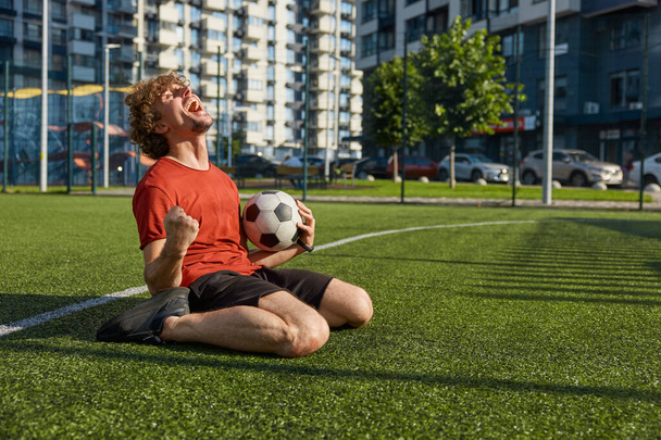 Portrait of euphoric soccer player screaming clenching fist while sitting on grass of outdoor stadium. Male footballer making winner gesture and celebrating victory of his team - Photo, Image