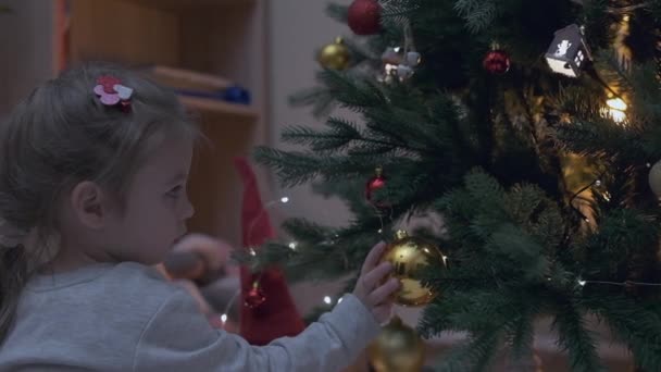Pieni viehättävä tyttö mekko ripustaa kiiltävä pallo joulukuusi. Lapsi on koristelu joulukuusi kauniissa olohuoneessa. Onnellisen perheloman käsite Uusi vuosi. Korkea - Materiaali, video