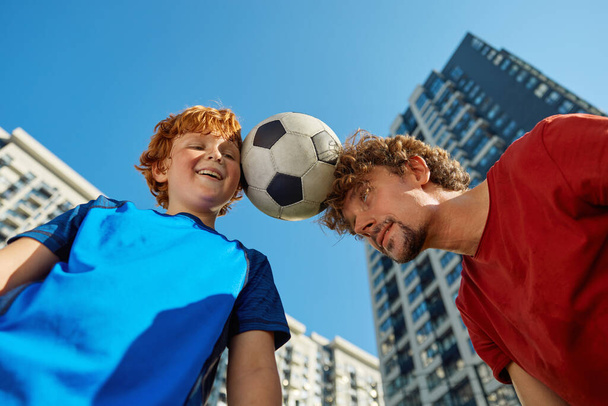 Closeup of adult man and boy child holding soccer ball between forehead view from bottom over city skyscraper background. Family competition, combat son against father - Photo, Image