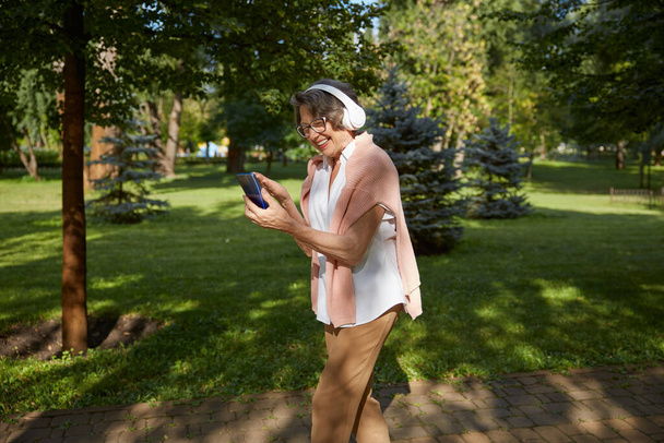 Vecchia anziana in cuffia che fa videochiamate online su smartphone utilizzando la connessione internet wireless con parenti o amici mentre cammina nel parco - Foto, immagini