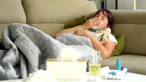 Una mujer de mediana edad se acuesta tosiendo, cubriéndose la boca, usando una servilleta de papel. Una mujer tiene un ataque de tos. Enfermedad y mala salud en el hogar - Imágenes, Vídeo