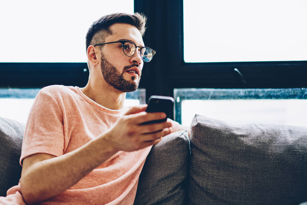 Caucasian bearded man in stylish optical spectacles resting on apartment sofa with smartphone gadget in hand and thinking about fresh interesting ideas for next publication in social network - 写真・画像