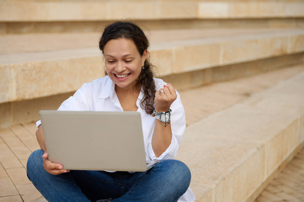 Gelukkig Latina zakenvrouw klemmen vuisten tijdens het online werken op laptop, buiten zitten op stappen. Doelstellingen bereikt. Succes. Mensen. Carrière en rekrutering. Online communicatie en werken op afstand - Foto, afbeelding