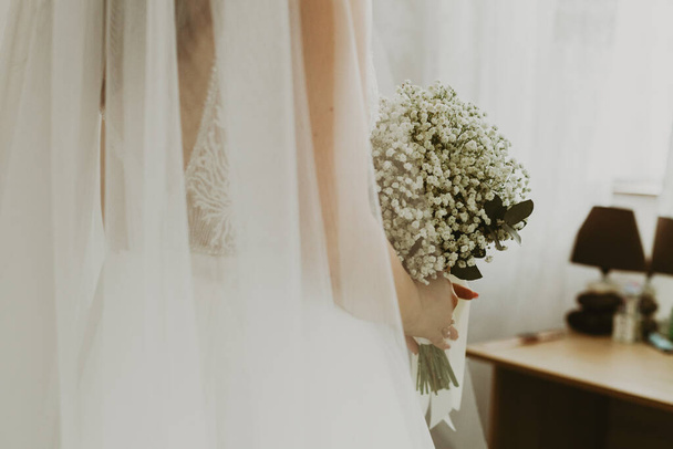 Portrét jedné krásné mladé kavkazské nevěsty držící během dne v místnosti kytici bílých boutonniér, pohled zdola, zblízka s výběrovým zaměřením. - Fotografie, Obrázek