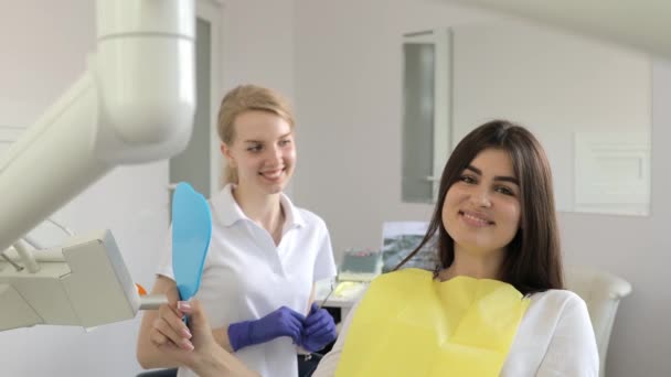 若い幸せな女性のクライアントは,歯科オフィスで微笑みながら鏡を見ています. 歯医者オフィスの鏡で歯をチェックする美しい若い女性は,完璧なまっすぐな白い歯を示しています. - 映像、動画