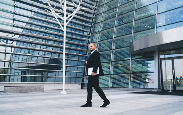 Боковой вид на бородатого бизнесмена в черном костюме, идущего перед стеклянной стеной современного офисного здания днем - Фото, изображение