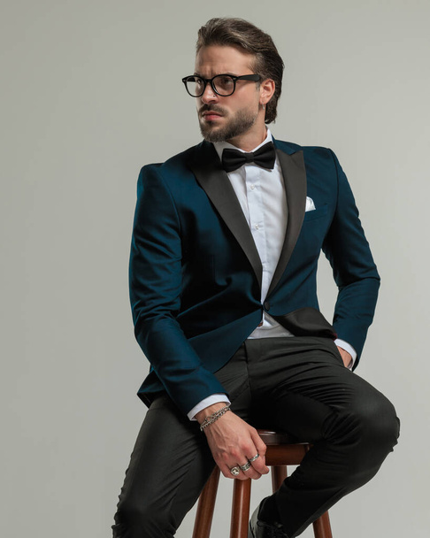 homem de moda barbudo com óculos olhando para o lado e segurando mão no bolso enquanto sentado na frente de fundo cinza  - Foto, Imagem