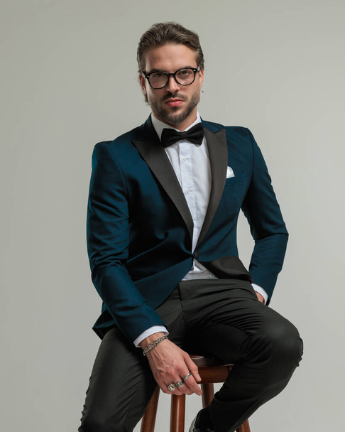 confident high class man in tuxedo with glasses sitting and posing with hand in pocket in front of grey background - Photo, Image