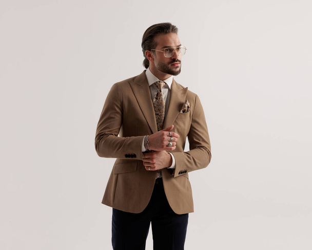 ritratto di uomo attraente in abito marrone con occhiali che guardano a lato e regolare vestito mentre si cammina di fronte a sfondo grigio - Foto, immagini