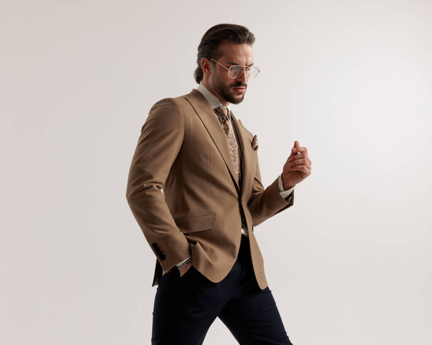 elegantní mladý muž s brýlemi v hnědém obleku drží ruku v kapse, dívá se dolů a pózuje chladným způsobem na šedém pozadí - Fotografie, Obrázek