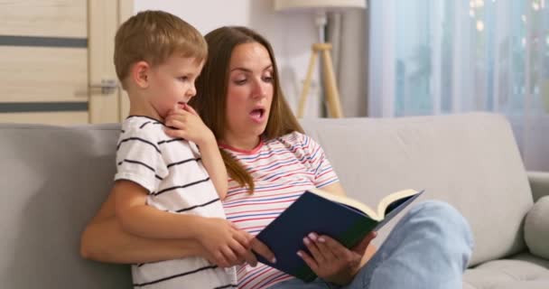 Matka i syn czytają książkę, siedzą na wygodnej sofie w salonie, spędzają czas z rodziną. Wysokiej jakości materiał 4k - Materiał filmowy, wideo