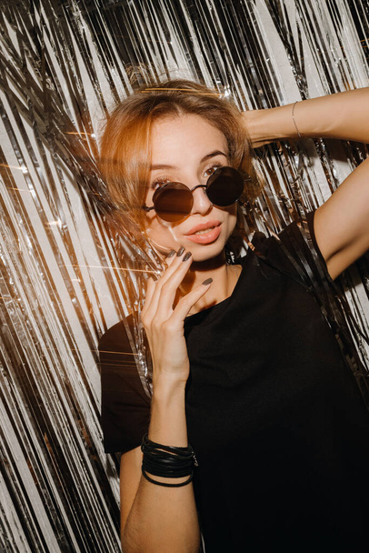 Retrato de revista vertical da menina europeia vestindo óculos redondos contra cortina cinza brilhante segurando a mão no cabelo e olhando para a câmera - Foto, Imagem