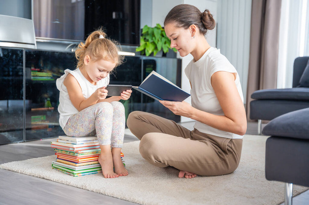 Klein meisje zit op een stapel kinderboeken en gebruikt haar smartphone terwijl moeder een boek leest. Hoge kwaliteit foto - Foto, afbeelding