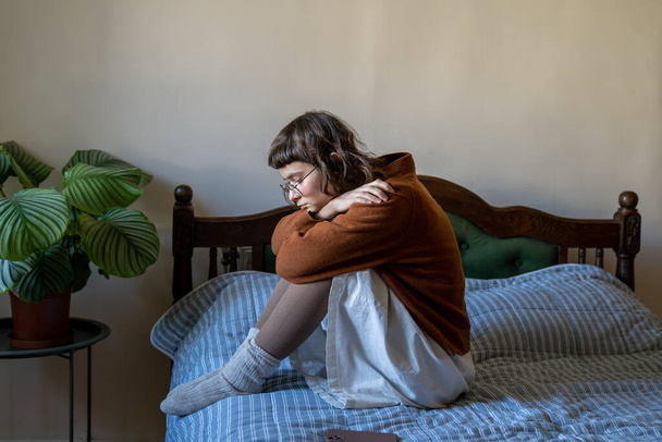Апатичный безэмоциональный интроверт-подросток, сидящий на кровати, обнимающий колени в одиночестве дома. Дружба, одиночество, депрессия, отсутствие мотивации, потеря жизненного интереса, концепция самоуничтожения - Фото, изображение