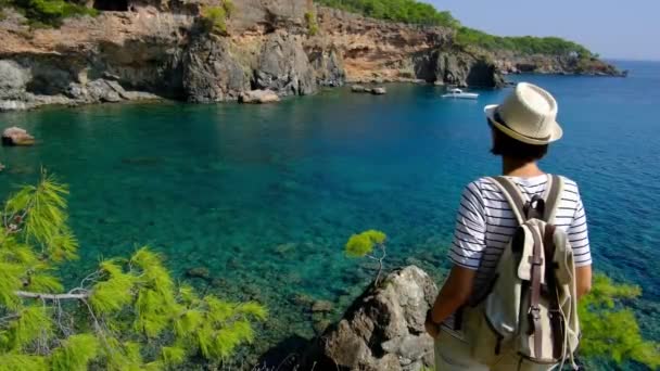 Tourist sightseeing on travel holiday. Back view. A female tourist with backpack walks along path along sea through forest. summertime travel in Antalya Turkey - Filmmaterial, Video