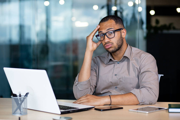 Hombre frustrado y triste en el lugar de trabajo leyendo el mensaje en línea de la computadora portátil, hombre de negocios sentado dentro de la oficina en el lugar de trabajo, desesperado infeliz, tiene malas noticias, bancarrota, despido. - Foto, imagen