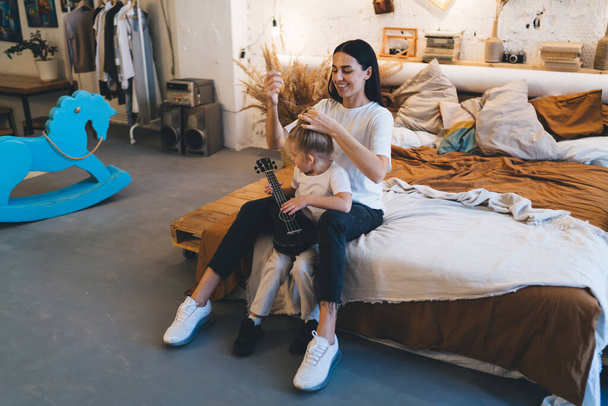 Positive young female mother fixing hair of daughter in ponytail while having fun and spending free time sitting on bed with cute girl in decorated bedroom - 写真・画像