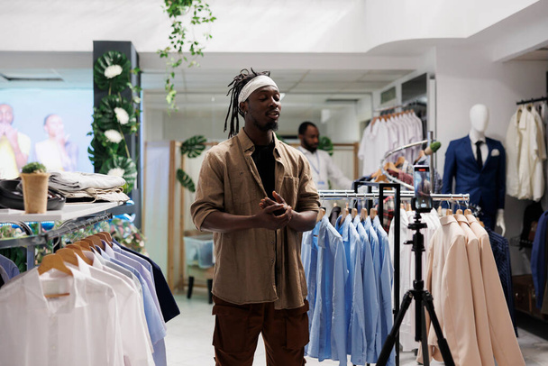 Influencer afroamericano che filma video in diretta su smartphone per mostrare i vestiti in negozio, parlando con i follower sulla fotocamera. Blogger che promuove la collezione di abbigliamento per i social media - Foto, immagini