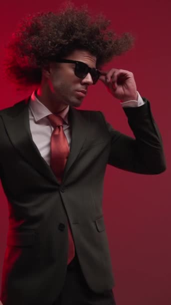 σέξι σγουρά μαλλιά άνθρωπος με γυαλιά ηλίου που αναζητούν στο πλάι, στερέωση γυαλιά ηλίου και αισθησιακά αγγίζοντας τα χείλη σε γκρι φόντο - Πλάνα, βίντεο