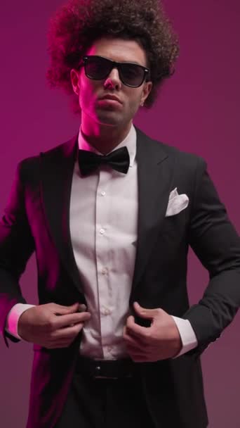 Projektionsvideo eines eleganten jungen Bräutigams mit Sonnenbrille, der nach vorne blickt, sich anpasst und schwarzen Smoking knöpft, bevor er auf buntem Hintergrund zur Seite schaut - Filmmaterial, Video