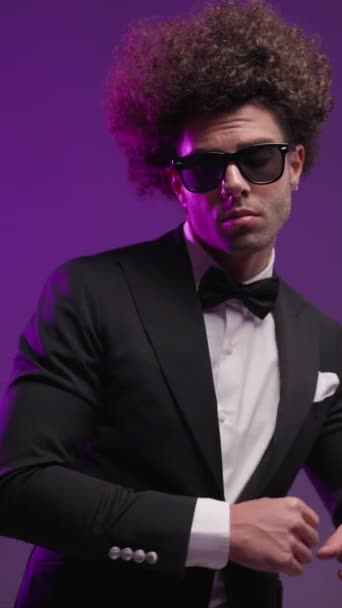 vídeo vertical del novio de moda fresco con gafas de sol mirando al lado y bailando, ajustando el traje y tomándose de la mano en bolsillos sobre un fondo colorido - Metraje, vídeo