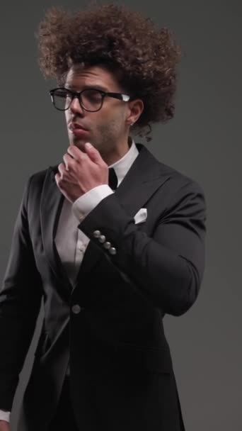 proyecto de vídeo de un joven elegante hombre con gafas de vista con esmoquin negro y rostro conmovedor mientras posa delante de fondo gris - Imágenes, Vídeo
