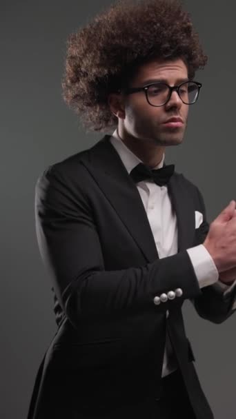 lockiges Haar Geschäftsmann mit Brille in schwarzem Smoking Blick zur Seite, Handflächen reiben und selbstbewusst vor grauem Hintergrund - Filmmaterial, Video