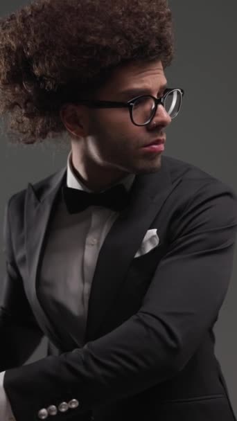 элегантный молодой человек в очках глядя в сторону, массируя ладони и регулируя черный галстук-бабочку перед серым фоном - Кадры, видео