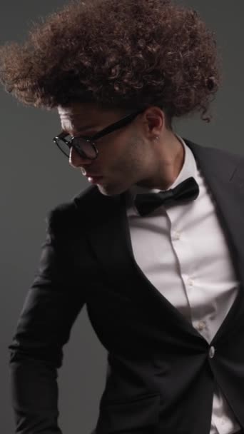 vertikales Video eines eleganten Bräutigams mit Brille, der die Hände in den Taschen hält, zur Seite schaut, Tux öffnet, Handflächen reibt und Kinn vor grauem Hintergrund berührt - Filmmaterial, Video