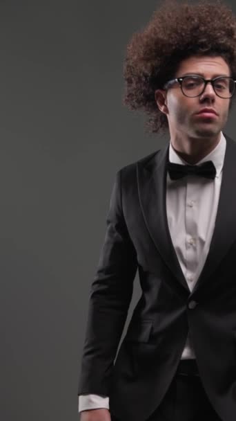 elegante hombre de moda con gafas cogidas de la mano en los bolsillos y mirando a un lado antes de tocar la cara sobre fondo gris - Metraje, vídeo
