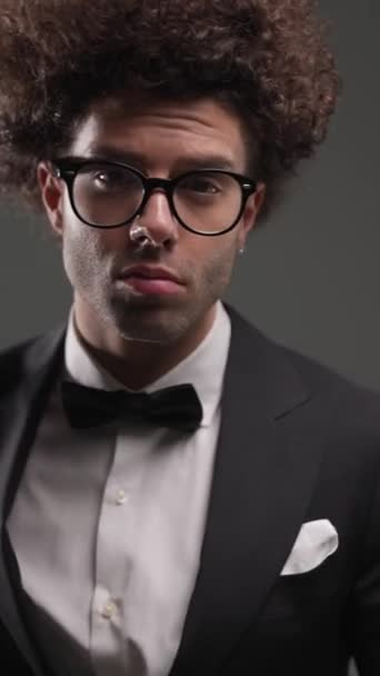 primer plano vídeo de elegante joven novio en esmoquin negro mirando hacia arriba y la organización de las gafas, mirando a un lado sobre fondo gris - Imágenes, Vídeo