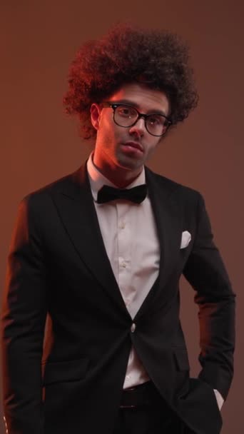 happy elegant groom with eyeglasses in black tuxedo holding hand in pocket and walking away on grey background - Footage, Video