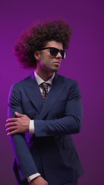 homem confiante com cabelo encaracolado usando óculos de sol, de mãos dadas em bolsos, ajustando o terno e segurando a mão atrás do pescoço enquanto sorri na frente de fundo colorido - Filmagem, Vídeo